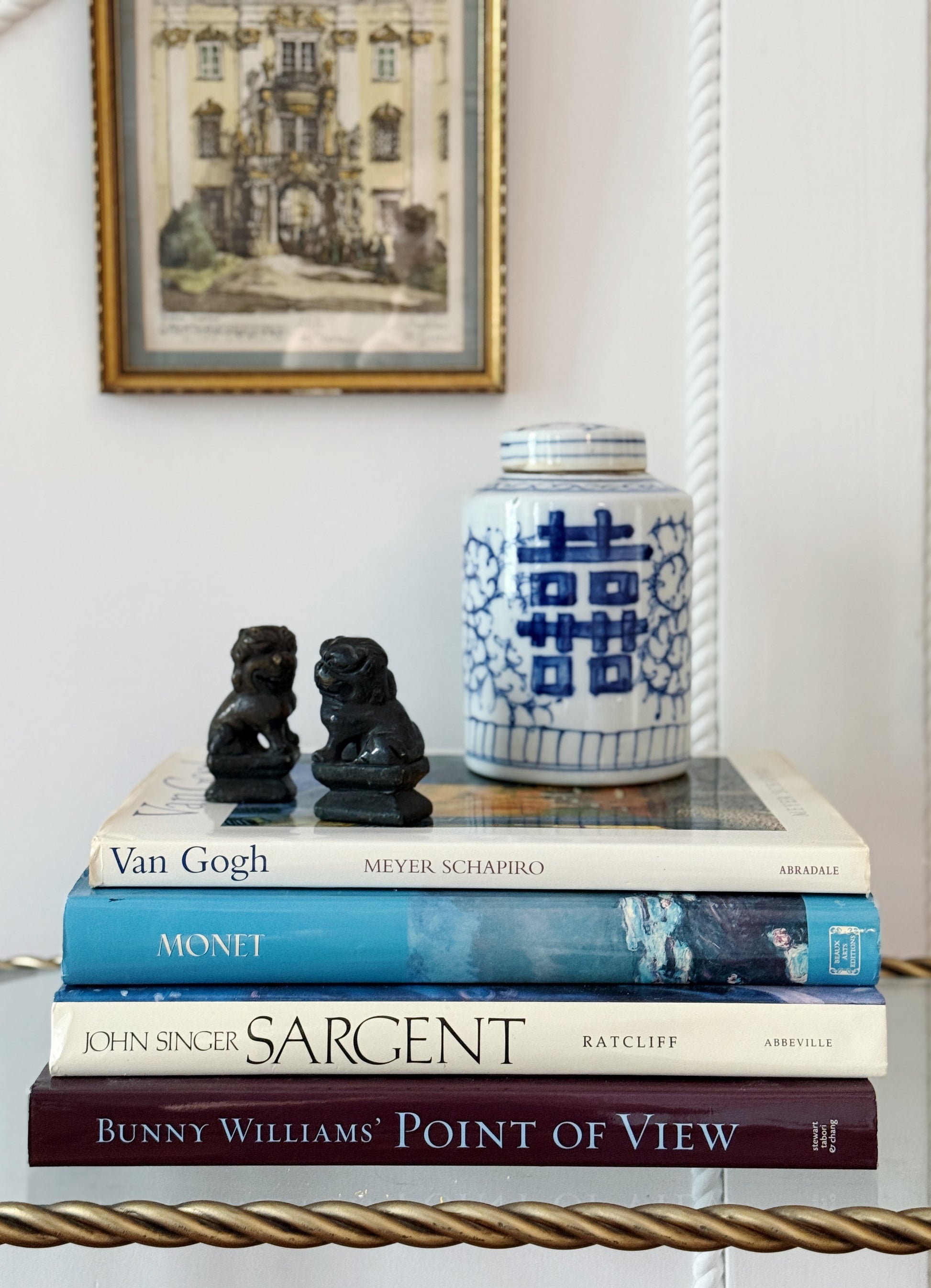 Vintage coffee table books with a blue and white ginger jar and marble foo dogs under a vintage piece of art