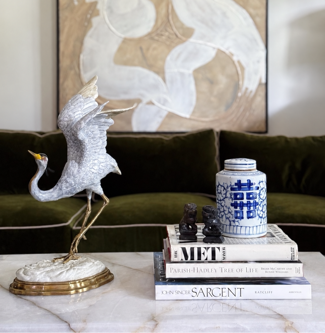 A porcelain crane sculpture styled with vintage coffee table books and blue and white chinoiserie porcelain in a green velvet living room