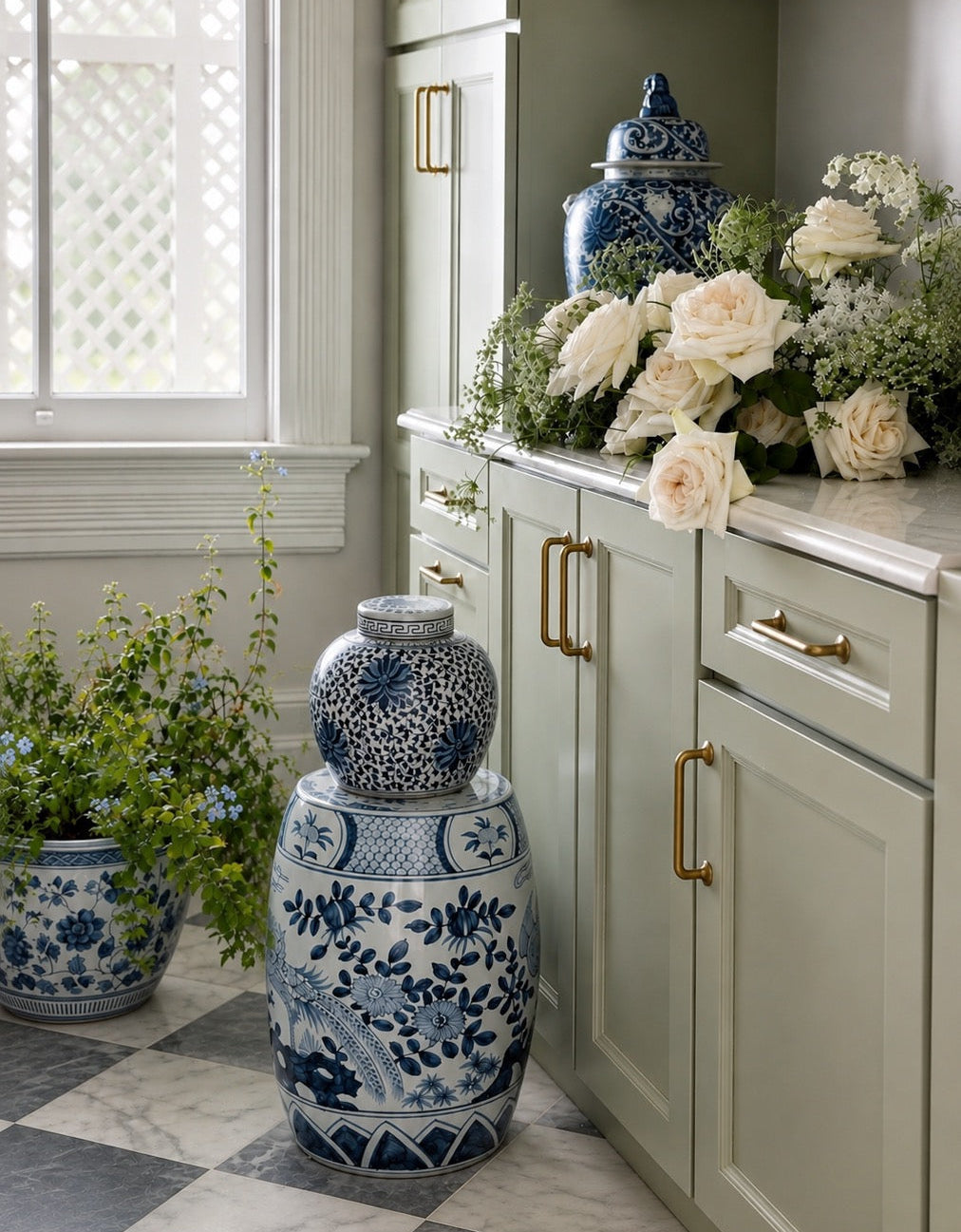 A styled interior corner with pale green cabinets, brass handles, a window with a bamboo blind, and blue-and-white porcelain garden vases and floral arrangements on the counter and floor.